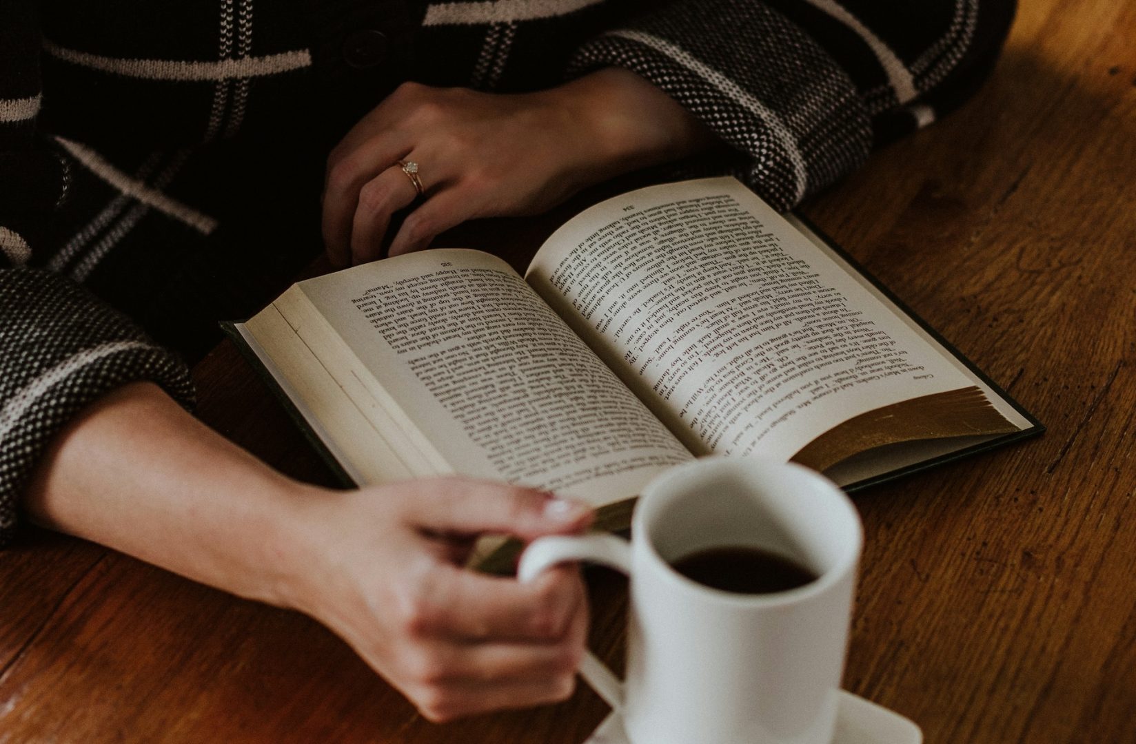 A person sips coffee while engrossed in an open book, the soft glow of a wooden table setting the stage—much like crafting a cozy design in an indie author website.
