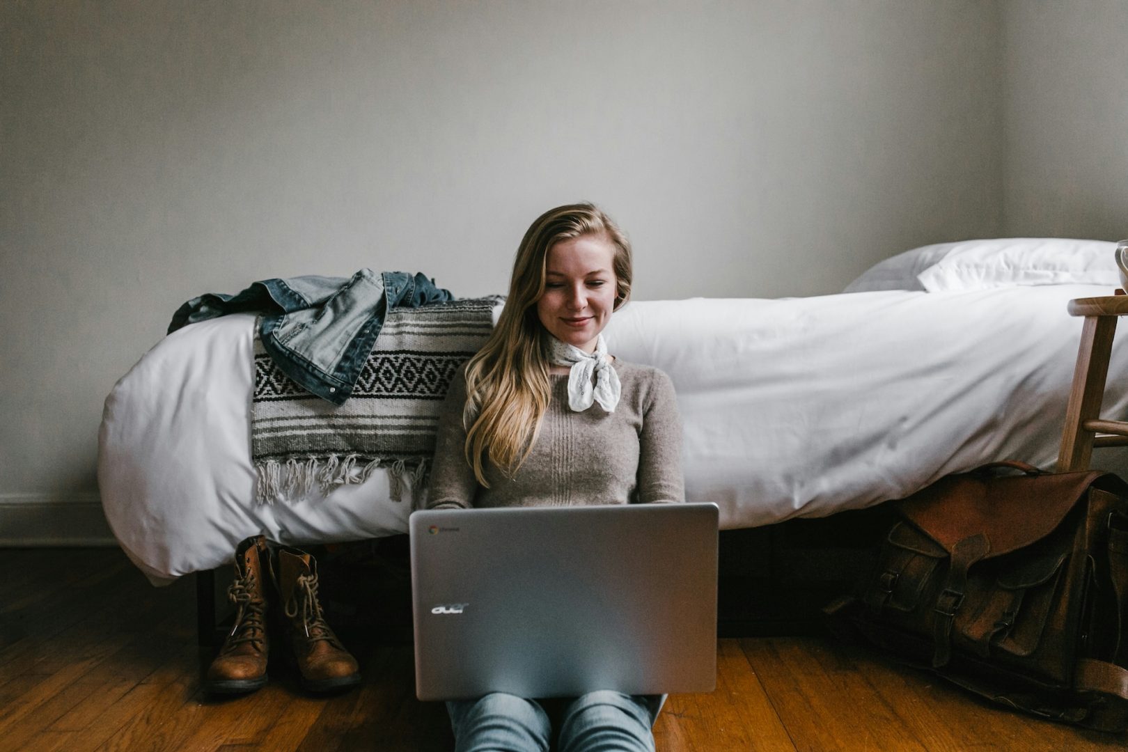 A digital-first enthusiast sits cross-legged on the floor, immersed in their laptop, with a bed and scattered clothes providing a casual backdrop.